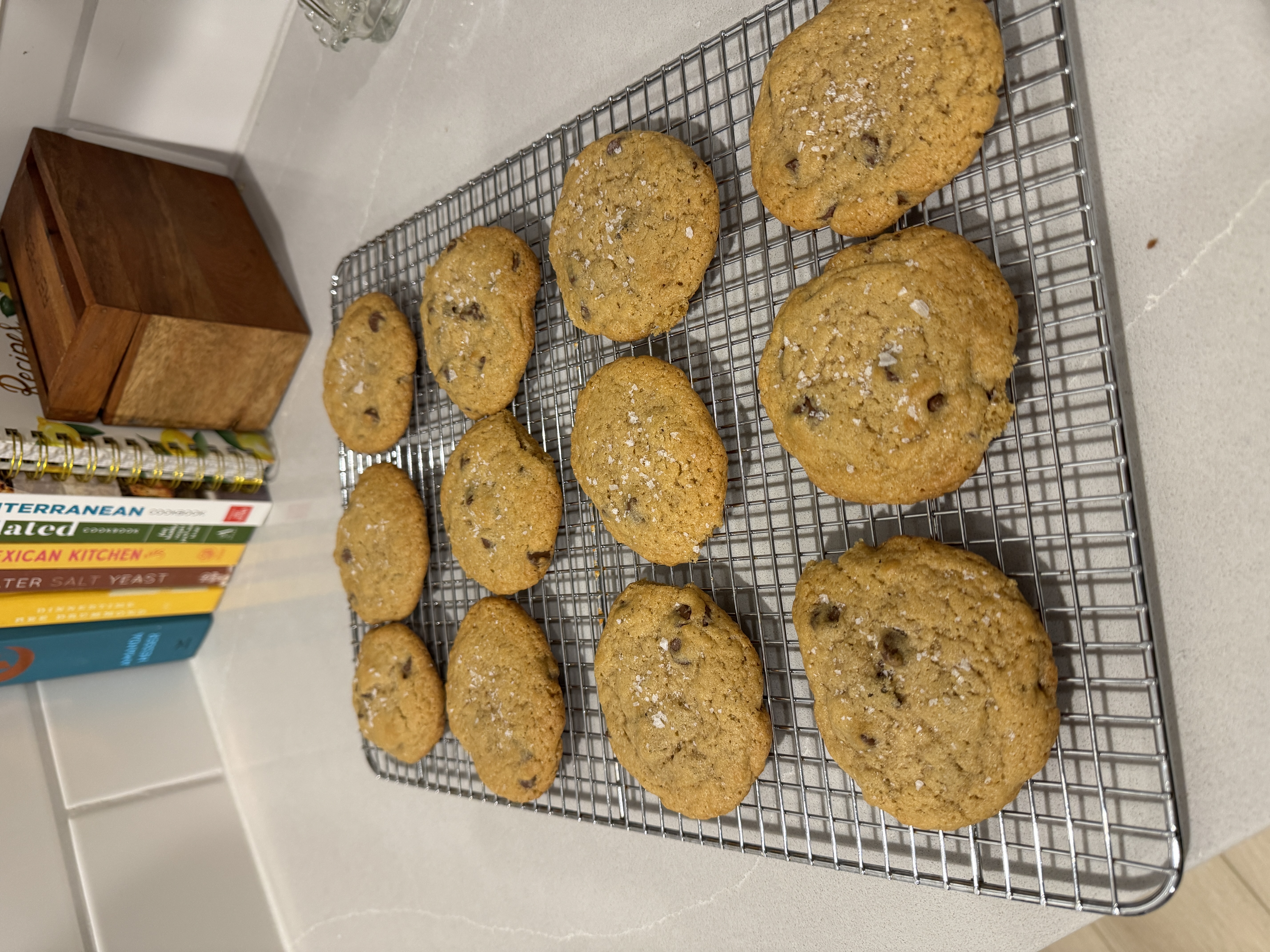 A tray of large rustic chocolate chip cookies cooling on a wire rack, flecked with sea salt.