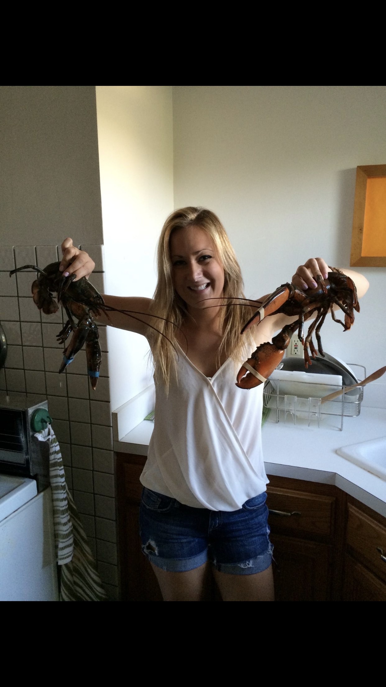 Sara laughing in her kitchen, holding two live lobsters, cookbooks stacked behind her.